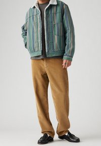 Chaqueta de mezclilla a rayas verdes y azules con un cuello de sherpa blanco, combinada con pantalones de pana beige y mocasines negros. Presenta un bolsillo en el pecho.