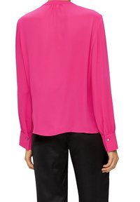 En kvinna ses bakifrån, iklädd en långärmad, klar rosa blus med en samlad hals och knäppningar vid ärmsluten, tillsammans med svarta byxor.