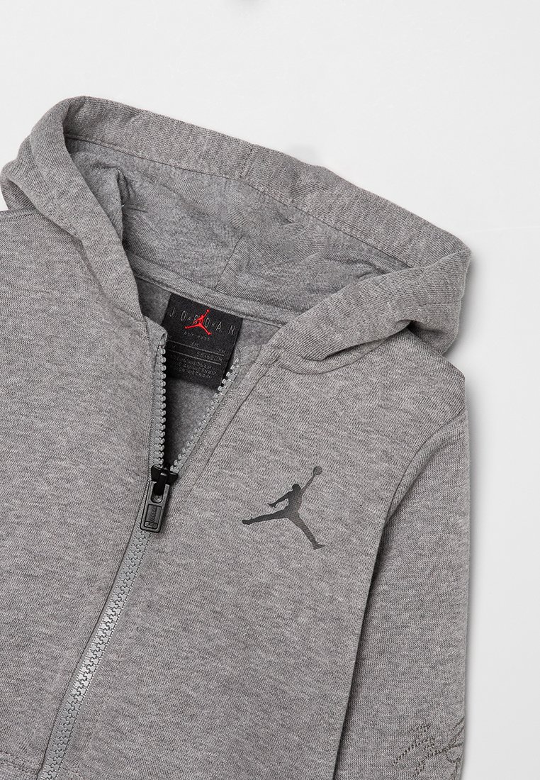 Hodie gris zippé en tissu doux, avec une capuche, un logo noir sur la poitrine et des détails de couture visibles le long de la fermeture éclair.