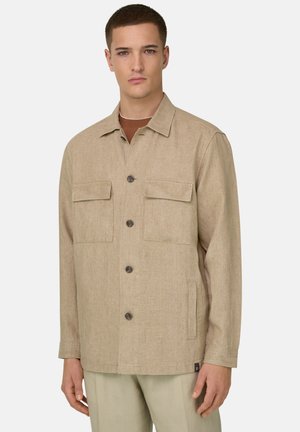 Jonge man draagt een beige jas met knopen, klepzakken op de borst en zijzakken met openingen, staand tegen een effen lichte achtergrond.