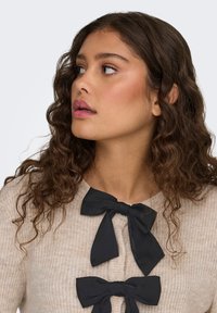 Beige neulottu neuletakki, jossa kolme mustaa nauhabussia edessä. Kangas on kuvioitua ja pääntie on pyöreä.