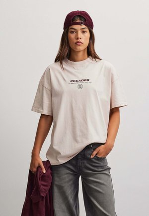 Jonge vrouw draagt een lichtbeige Pegador T-shirt en een bordeauxrode pet achterstevoren, houdt een bordeauxrode jas vast, met één hand in de zak van een grijze spijkerbroek.