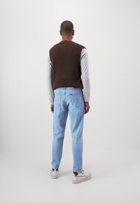 Maglione marrone senza maniche sopra una camicia grigia a maniche lunghe, abbinato a jeans blu chiaro. Sneakers bianche con dettagli marroni. Sfondo minimalista.
