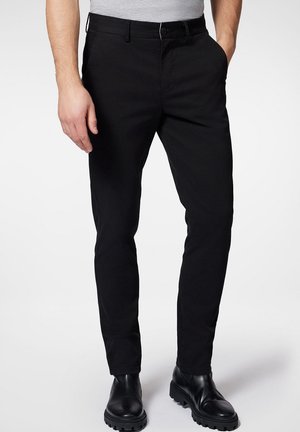 Uomo che indossa pantaloni neri aderenti con tasche frontali e stivali neri in pelle, in piedi su uno sfondo chiaro e semplice.