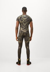Man facing away wearing black and gold futuristic skull-patterned athletic shirt and leggings, black sports shoes with yellow accents.