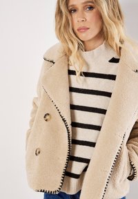 Beige bontjas met zwarte stiksels, dubbelzijdig ontwerp, met grote knopen. Draag het over een gestreept trui in crème en zwart.