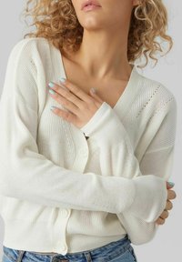 Cardigan en tricot blanc avec un col en V, doté d'une fermeture à boutons, de poignets côtelés et d'un motif texturé. Tissu doux et léger.