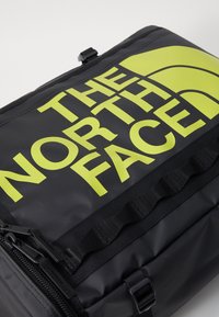 Sac à dos noir présentant un grand logo jaune audacieux. Le design comprend un tissu texturé et des compartiments zippés, avec des accents en métal.