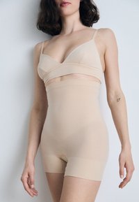 Béžový tvarujúci body s hladkým, elastickým materiálom. Zaujímavý dizajn s prekríženým predným dielom a krátkym pokrytím nôh, ktorý zabezpečuje bezšvový vzhľad.