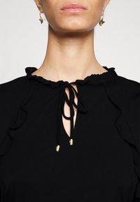 Svart topp med volangkrage och knytning, synliga gyllene accenter och en nyckelhålsdetalj i mitten. Slät yta.