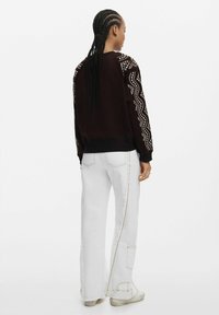 Hanorac maro cu broderie geometrică albă pe umeri, asortat cu pantaloni albi cu croi larg, care au cusături aurii și un buzunar tip cargo.