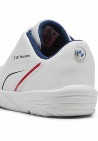 Chaussure de sport blanche avec des accents marine, des lignes rouges et noires, une tige texturée et une semelle en caoutchouc. Présente le logo BMW au niveau du talon et de la marque sur le côté.