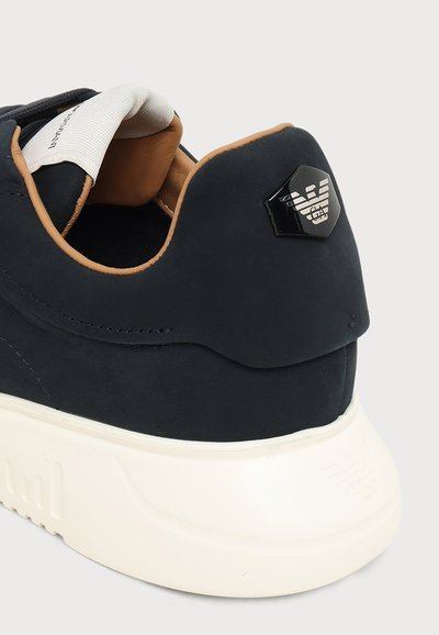 Talons de baskets noirs avec semelle blanche, doublure intérieure beige et un badge logo hexagonal noir à l'arrière.
