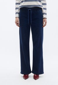 Pantalones anchos azul marino con cintura elástica y cordón. El material parece suave con una textura lisa, combinados con zapatos rojos brillantes.