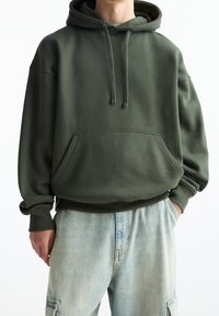 Felpa con cappuccio verde oliva con tasca frontale e coulisse, realizzata in tessuto morbido, indossata sopra pantaloni di denim azzurro chiaro.