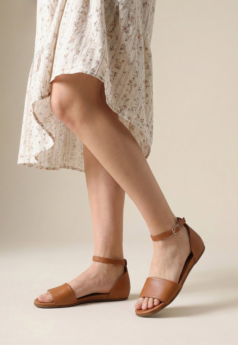 Gekruiste benen met tankleurige enkelsandalen met bandje en een crème bloemen hoge-lage rok tegen een lichte neutrale achtergrond.