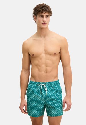 Uomo senza maglietta con capelli ricci che indossa pantaloncini da bagno turchesi a motivo con coulisse bianca, in piedi contro uno sfondo chiaro uniforme.