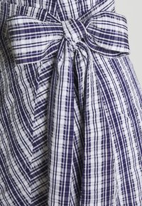 Lila och vit randig tyg med en knuten rosett detalj i midjan, har en texturerad, skrynklig yta och varierande randbredder.