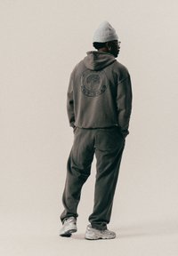 Osoba w szarej bluzie z kapturem i dresowych spodniach, z białą czapką beanie, w okularach, stojąca z rękami w kieszeniach, zwrócona plecami na jednolitym tle.