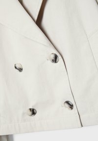 Nahaufnahme der Vorderseite eines beige-farbenen zweireihigen Mantels, die die Stoffstruktur, den Reverskragen und vier grau-marmorierte Knöpfe zeigt, die in zwei vertikalen Reihen angeordnet sind.