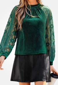 Groen fluwelen top met doorzichtige kant lange mouwen; heeft een hoge halslijn en wordt gecombineerd met een zwarte nepleren rok.