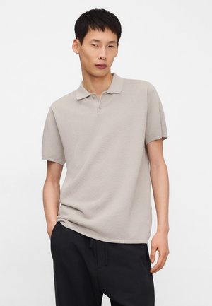 Młody mężczyzna w beżowej koszulce polo z krótkim rękawem i czarnych spodniach, stojący z jedną ręką w kieszeni na jednolitym tle.