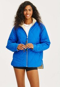 Modrá prošívaná bunda s krémovým interiérem, se zipem vpředu a kapsami. Modelka má na sobě bílé tričko a šortky, drží dekorativní korálky.