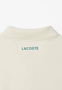 Gros plan de la zone arrière du cou d'un vêtement couleur crème avec un col côtelé et une étiquette rectangulaire au logo Lacoste centrée en dessous.