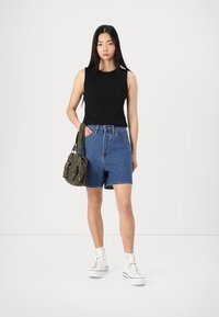 Black sleeveless top, high-waisted blue denim shorts, white platform sneakers, and green fabric handbag. Plain background.
