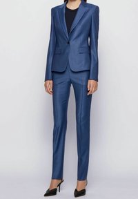 Costume bleu sur mesure avec un blazer structuré à revers crantés, deux poches avant et un pantalon droit, accompagné de talons noirs.
