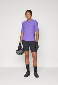 Chemise violette à manches courtes associée à un short noir. Gants noirs et casque tenus à la main, avec des chaussettes et des chaussures noires ajoutant du contraste.