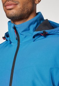Kék softshell dzseki magas gallérral, fekete béléssel, elülső cipzárral és piros díszüvegekkel a kapucniban és a nyak körül.