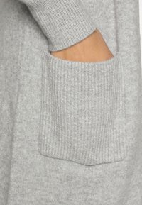 Hellgrauer Pullover mit weicher Textur, ausgestattet mit einem gerippten Bündchen und einer aufgesetzten Tasche auf der Vorderseite.