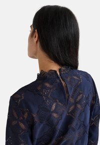 Blouse en dentelle bleu marine avec motifs floraux, col festonné et fermeture à boutons dans le dos. Détail en dentelle texturée sur toute la surface.
