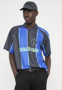 Μπλε και μαύρο κοντομάνικο πουκάμισο με κάθετες ρίγες, διαθέτει λαιμόκοψη σε σχήμα V, λογότυπο στο στήθος και αντίθετο πράσινο κείμενο.