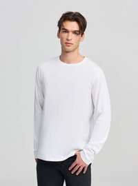 Biała longsleeve z miękkiego materiału o zaokrąglonym dekolcie, z luźnym fasonem i minimalnymi przeszyciami. Połączona z ciemnymi spodniami.