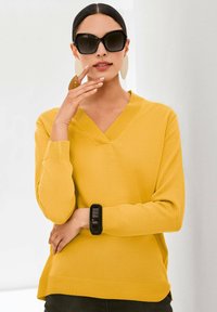 Gelber V-Ausschnitt-Pullover aus Strickmaterial mit gerippten Bündchen und Saum. Kombiniert mit übergroßen schwarzen Sonnenbrillen und einem schwarzen Armband.