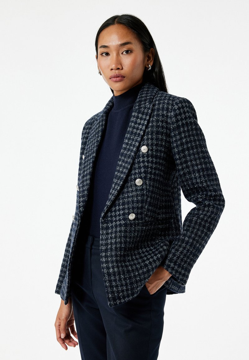 Marineblauwe textured blazer met een houndstooth patroon, dubbelbreasted ontwerp, zilveren knopen en opstaande revers. Gedragen over een donkere coltrui.