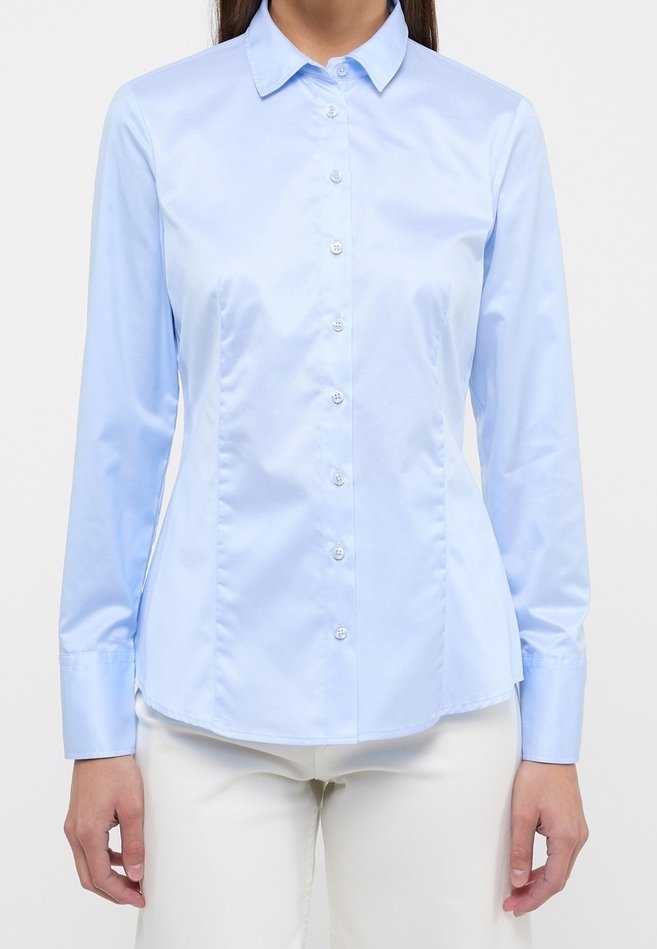 Femme portant une chemise bleu clair ajustée à manches longues boutonnée et un pantalon blanc, debout, les bras détendus le long du corps.