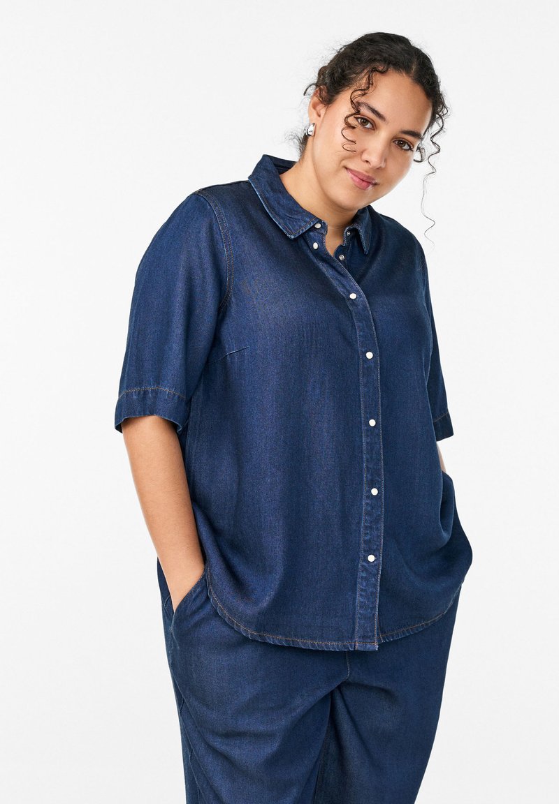 Nainen, jolla on kiharat hiukset ja yllään tummansininen farkkupaita ja siihen sopivat housut, kädet taskuissa, katsoo kameraan valkoista taustaa vasten.