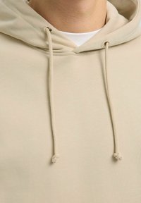 Felpa beige con una texture morbida, dotata di un cappuccio con lacci e due lacci simmetrici, abbinata a una camicia bianca sottostante.