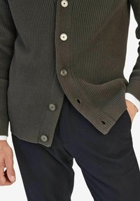 Mörkgrön ribbad stickad cardigan med fem stora knappar, sidofickor och en texturerad finish. Stylar med mörka byxor och vita ärmslut.