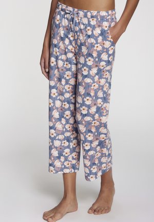 Woman wearing relaxed-fit, cropped pajama pants with blue background and pink and white floral pattern, one hand in pocket.