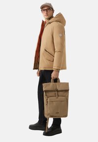 Beige Daunenjacke mit orangefarbenem Innenfutter, Reißverschlusstaschen und Kapuze. Das Model hält einen hellbraunen Rolltop-Rucksack mit einer Reißverschlusstasche auf der Vorderseite und Trägern.