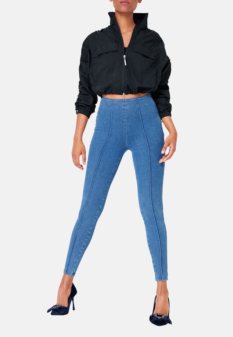 Zwarte cropped jas met een frontbijeen rits en twee zakken over blauwe high-waisted skinny jeans met contrasterende witte stiksels en een slanke vorm.