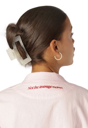 Rosa Hemd mit Kragen, mit roten gestickten Text „Nicht die durchschnittliche Mama“. Die Haare sind mit einer klaren, rechteckigen Haarklammer befestigt.