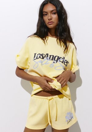 Jeune femme aux longs cheveux foncés porte un short jaune et un T-shirt ample assorti avec l’inscription « Los Angeles », ajustant l’ourlet sur un fond uni.