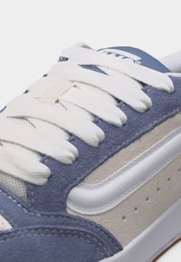 Κοντινή λήψη ενός μπλε και μπεζ αθλητικού παπουτσιού με λευκά κορδόνια και μια λευκή πλευρική λωρίδα πάνω σε ανοιχτό φόντο.