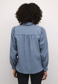 Personne aux cheveux bouclés portant un chemisier bleu à manches longues avec des épaules bouffantes et un pantalon noir, vue de dos sur un fond uni.