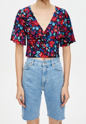 Person trägt eine schwarze, kurz geschnittene Bluse mit rotem und blauem Blumenprint und hellblaue, hoch taillierte Denim-Shorts mit ausgefransten Säumen.